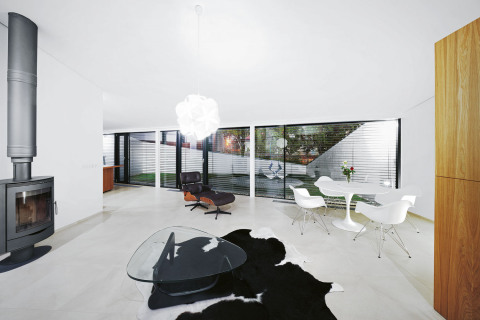 Společný obývací prostor si architekt přál naplnit světlem, proto zvolil bílé povrchy a čelní stěnu s velkoformátovým bezrámovým zasklením. Dominantou pokoje jsou válcovitá krbová kamna a slavné křeslo Lounge Chair od Charlese Eamese. Z jeho produkce pocházejí i bílá polokřesílka u jídelního stolu